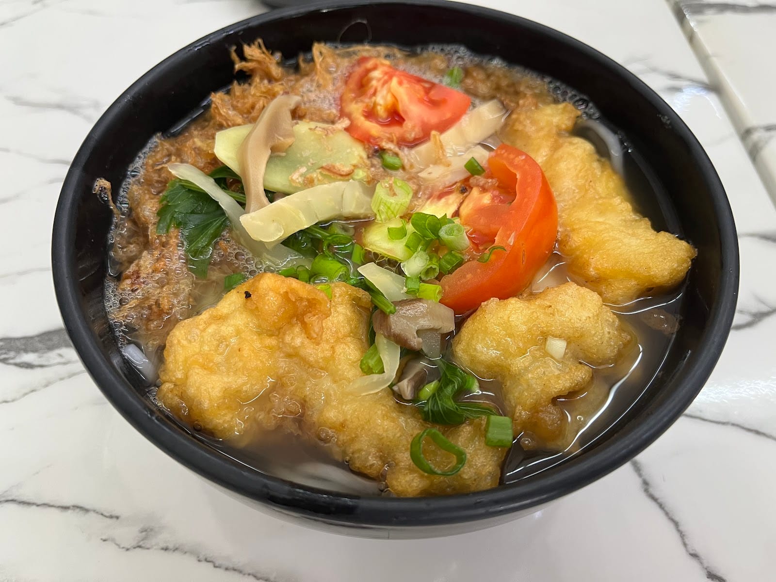 Fish mee hoon soup with battered fish, tomato and spring onion in a black bowl