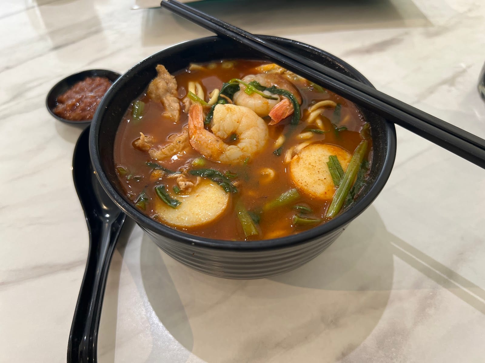 Prawn mee in a black bowl with prawns, fish cake slices and water spinach, chilli paste on the side
